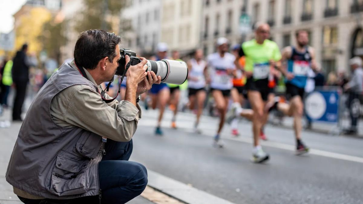 Photographe accroupi au bord de la route, appareil et téléobjectif pointés vers les coureurs d'un 10 km