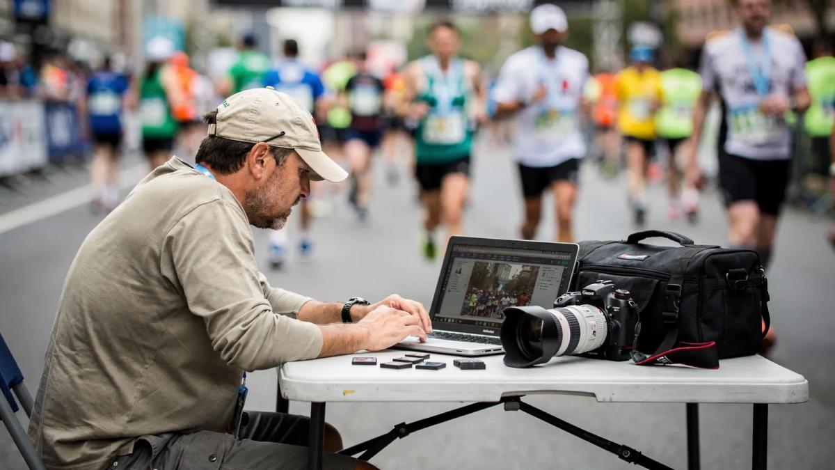 Photographe de course assis à une table près de l'arrivée, triant ses photos sur un ordinateur portable pendant que les coureurs passent